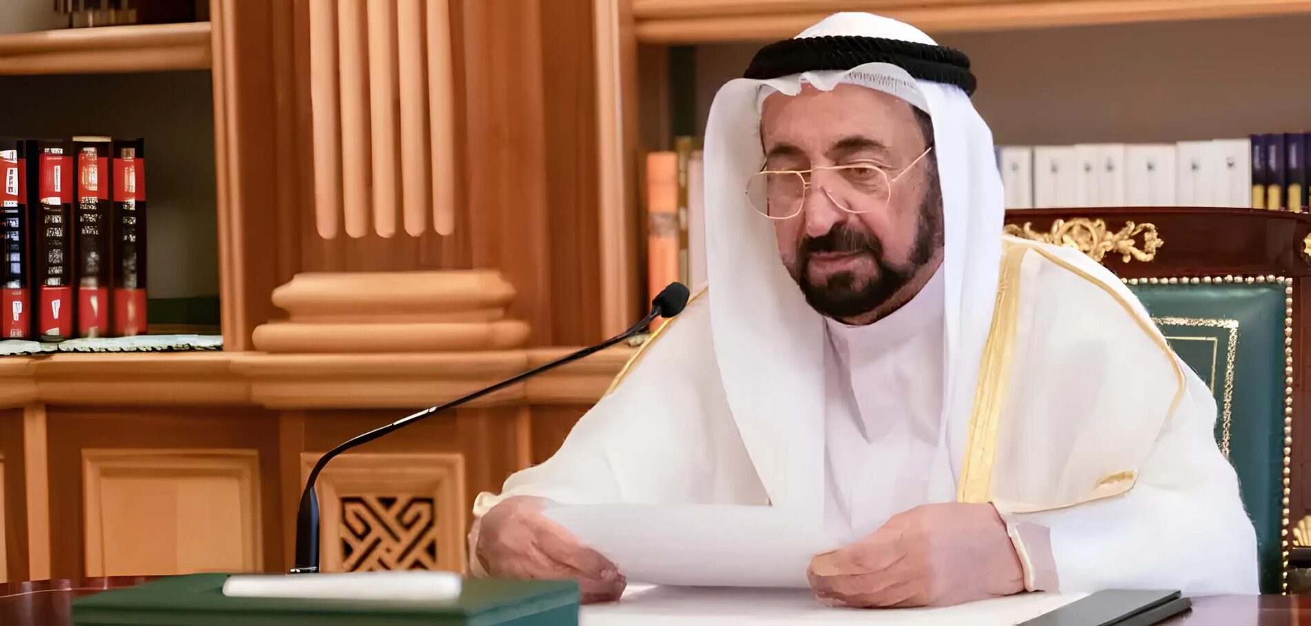 Sultan Muhammad Al Qasimi reading from a paper at a desk in a library setting, surrounded by bound volumes